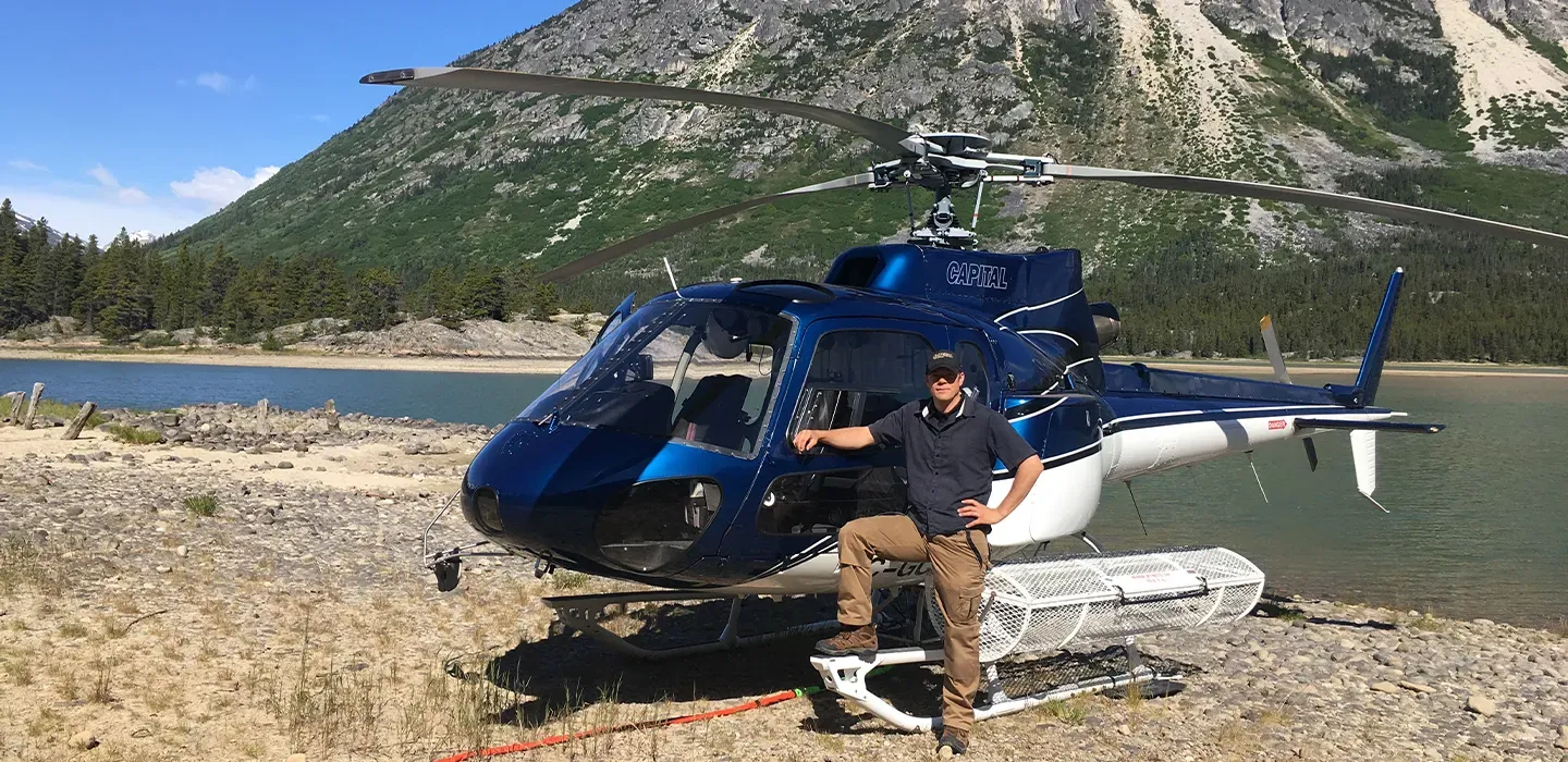 A Capital Helicopters pilot standing beside an AS350 helicopter parked on a rocky lakeshore beneath Yukon mountains